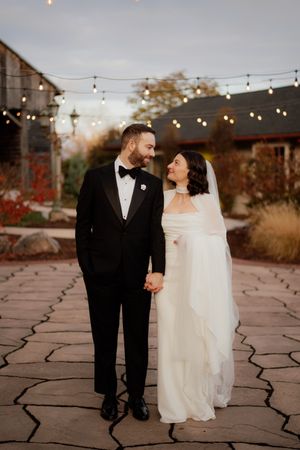 A couple walks together on a stone patio under string lights at dusk.