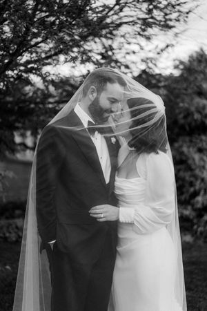 A romantic wedding moment captured in black and white as a couple shares an intimate moment under a flowing veil outdoors.