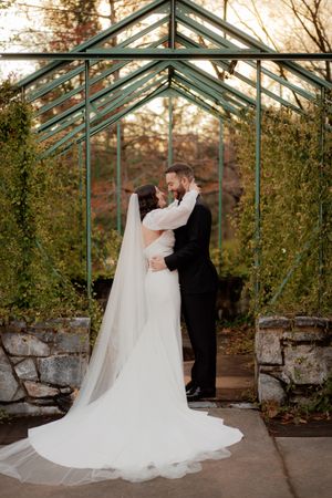 A romantic wedding couple shares an intimate moment in a glass greenhouse surrounded by autumn foliage.
