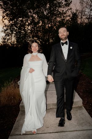A newlywed couple walks along a concrete path at sunset, with the bride wearing a flowing white dress.