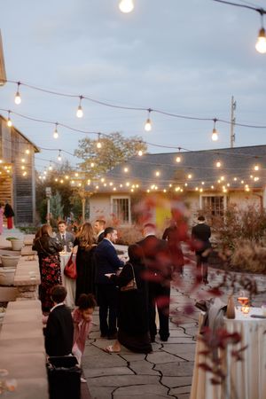 An outdoor wedding reception with string lights illuminating guests mingling on a stone patio at dusk.