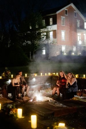 Group gathered around glowing campfire at night with luminaries and brick house in background.