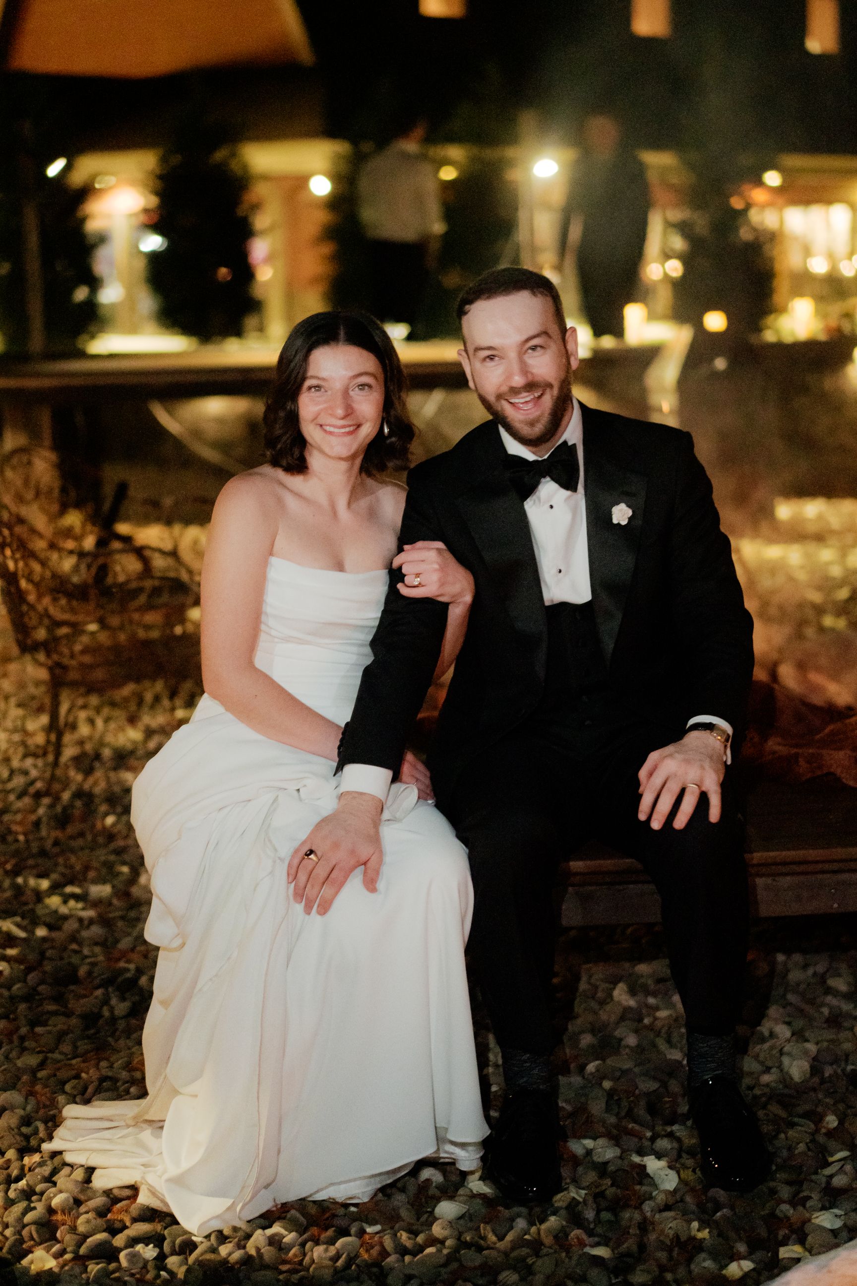 A romantic evening portrait of a couple sitting outdoors with twinkling lights in the background.
