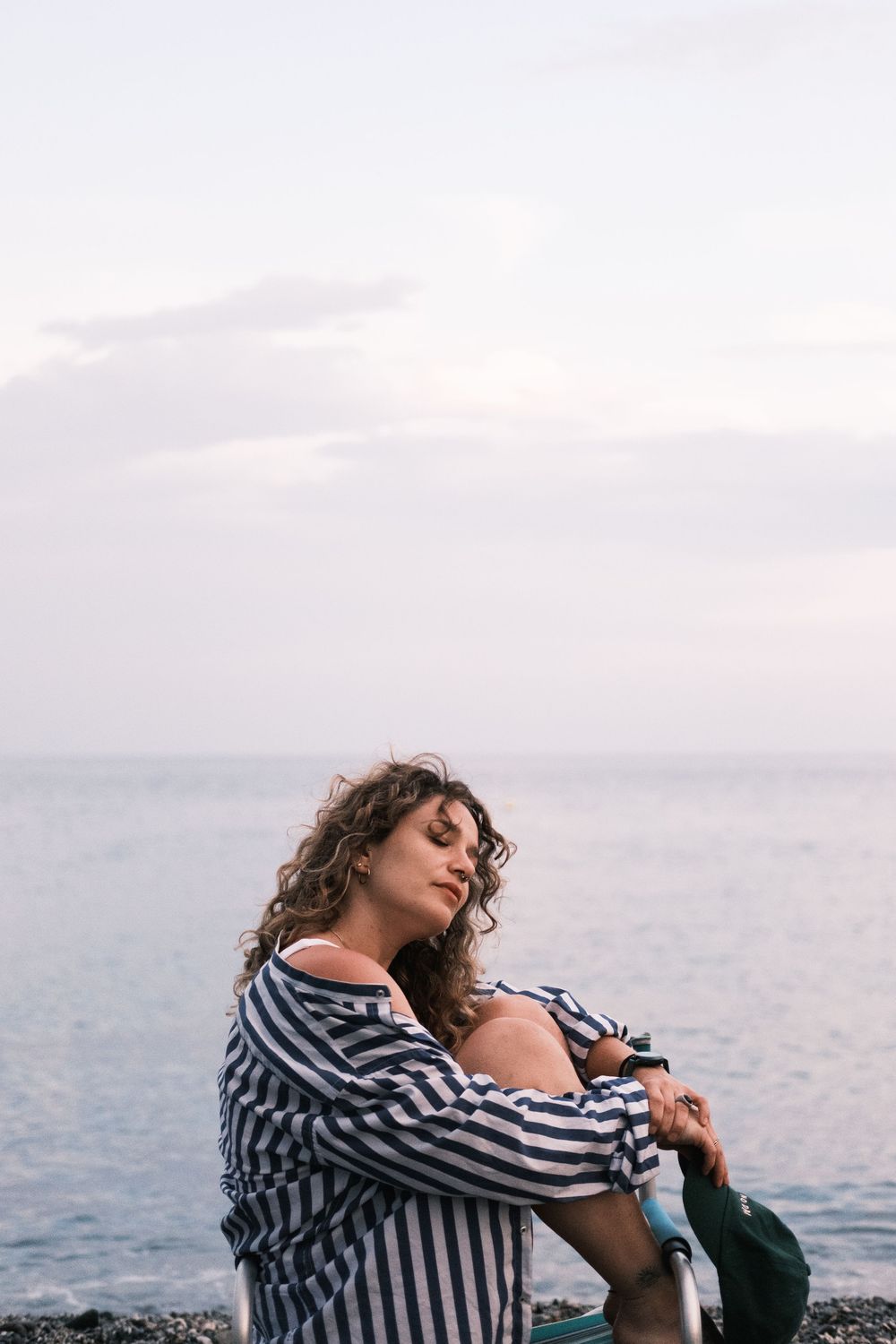 Person in striped shirt sits contemplatively by the ocean at sunset, creating a serene coastal scene.