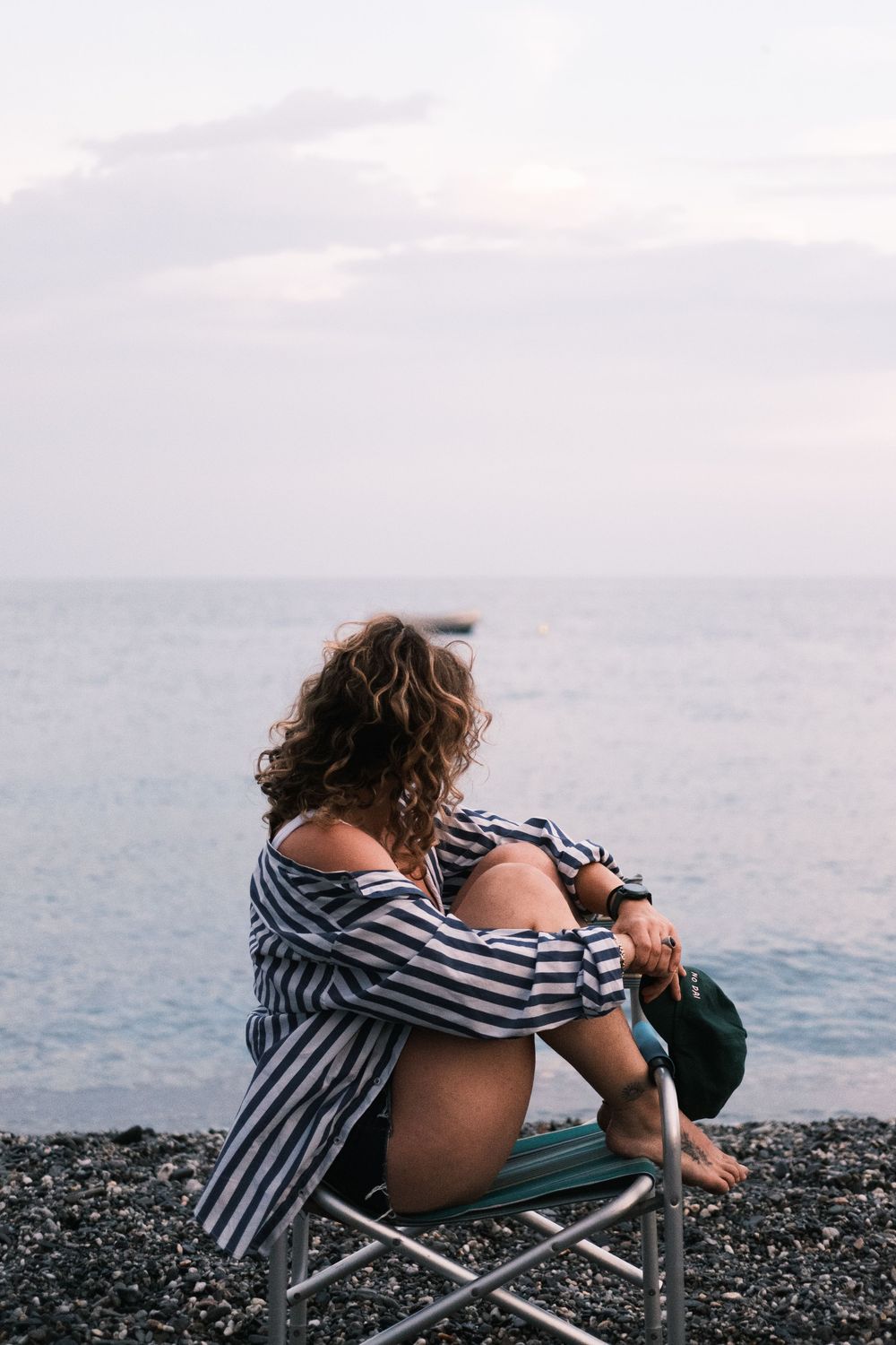 Person in striped shirt relaxes in beach chair on rocky shore overlooking calm ocean at sunset.