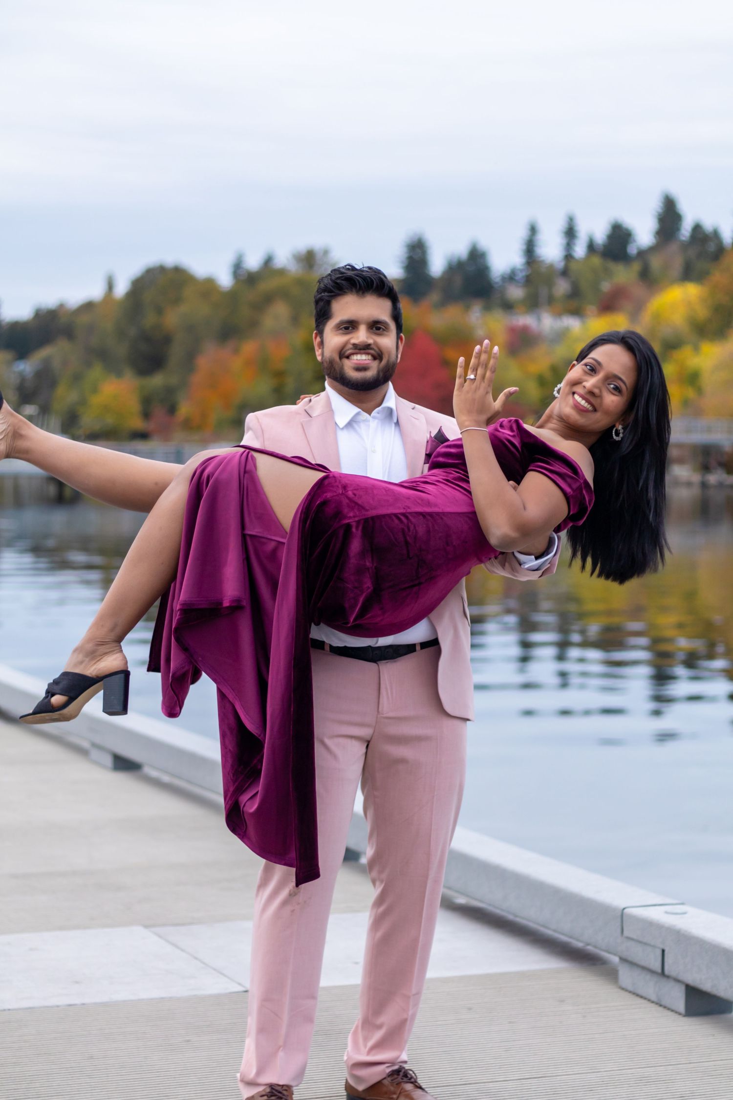 A man in a pink suit carries a woman in a magenta velvet dress by a waterfront with autumn foliage.