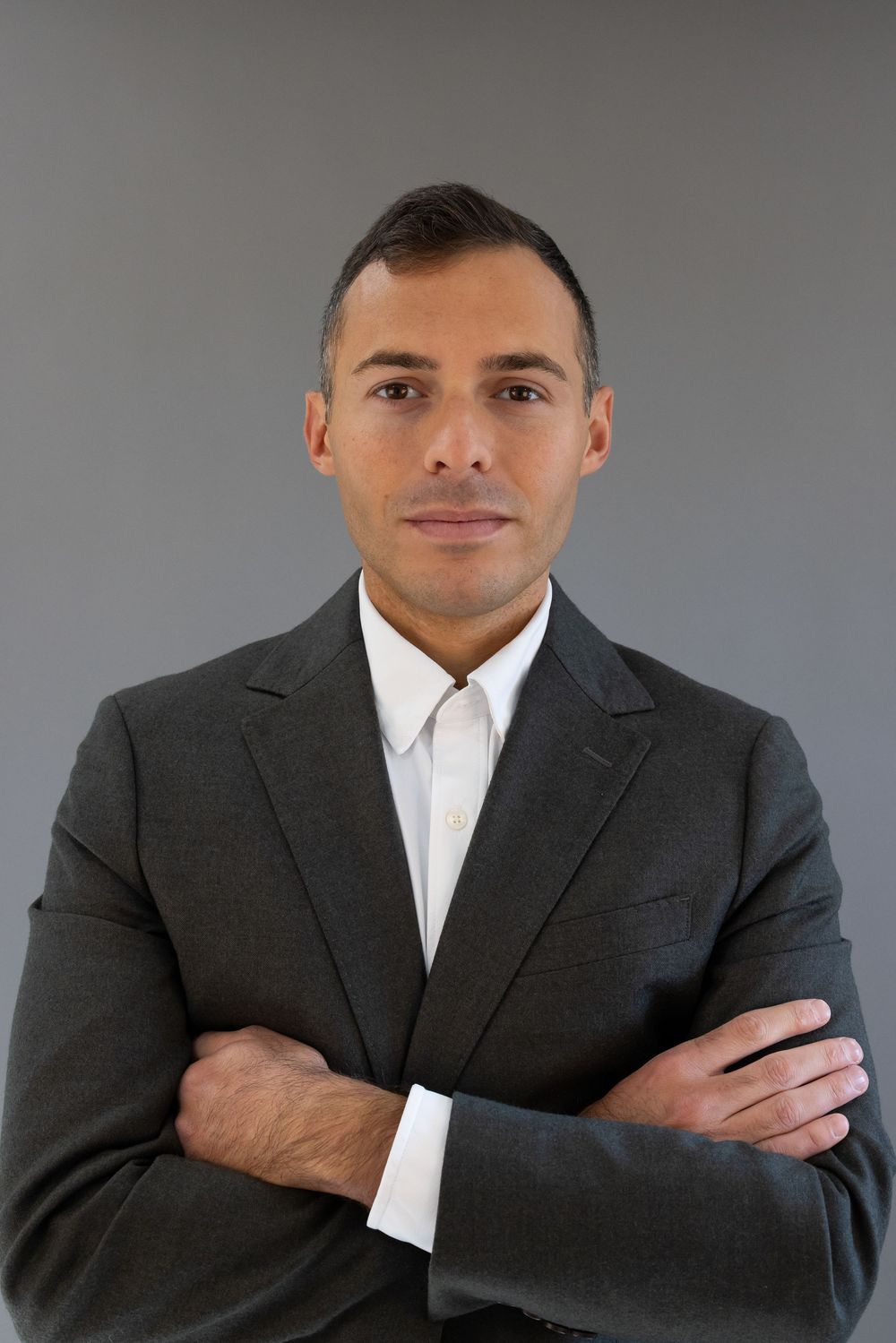 Professional wearing a dark gray suit and white shirt poses with crossed arms against a gray studio backdrop.