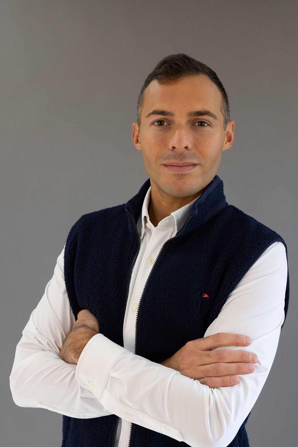 Professional headshot of someone wearing a navy vest over white dress shirt with arms crossed against grey background.
