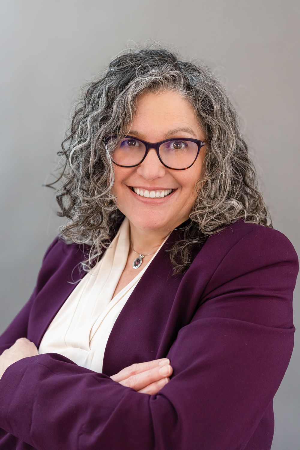 Professional headshots of someone wearing a purple blazer and white blouse with curly gray hair and glasses against gray background.