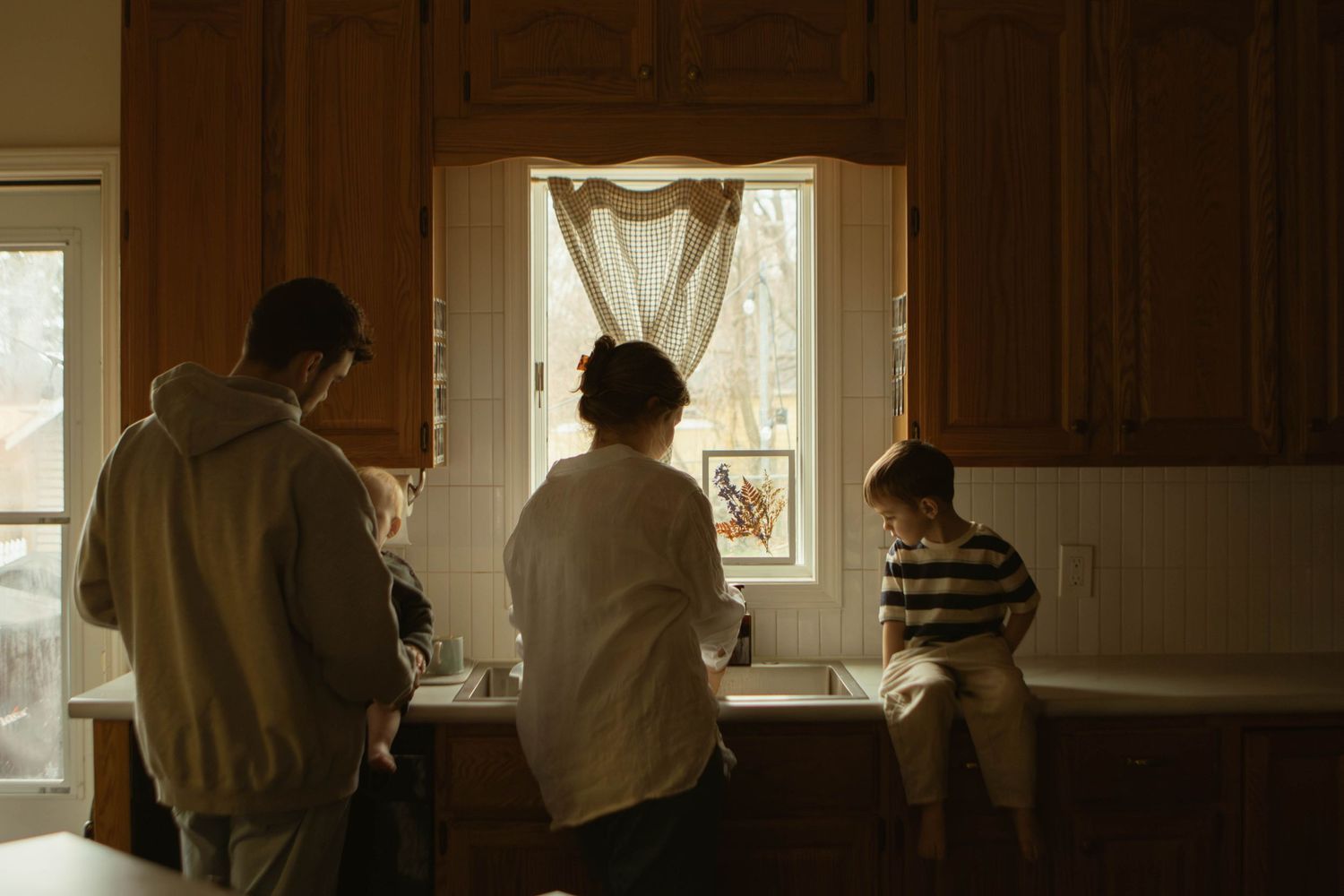 Family of four standing together at kitchen sink looking out sunlit window.