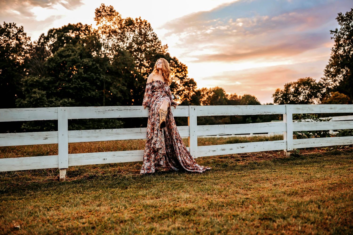 A rustic white fence stretches along a grassy hillside against a dramatic sunset sky with golden clouds.