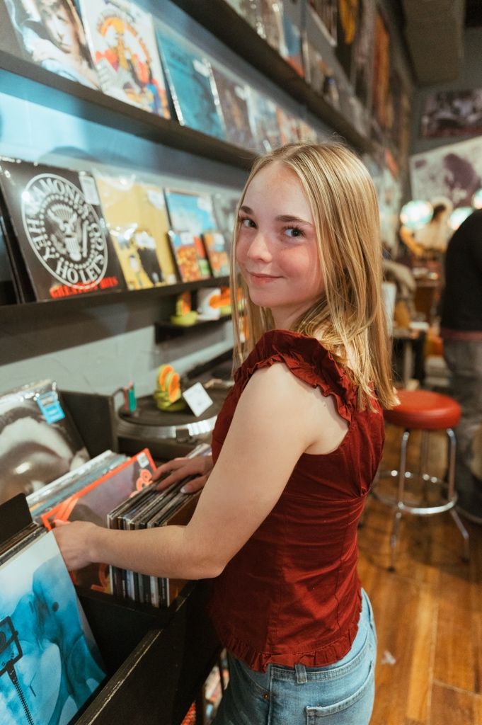 Person browsing vinyl records in a record store wearing a red velvet top and jeans.