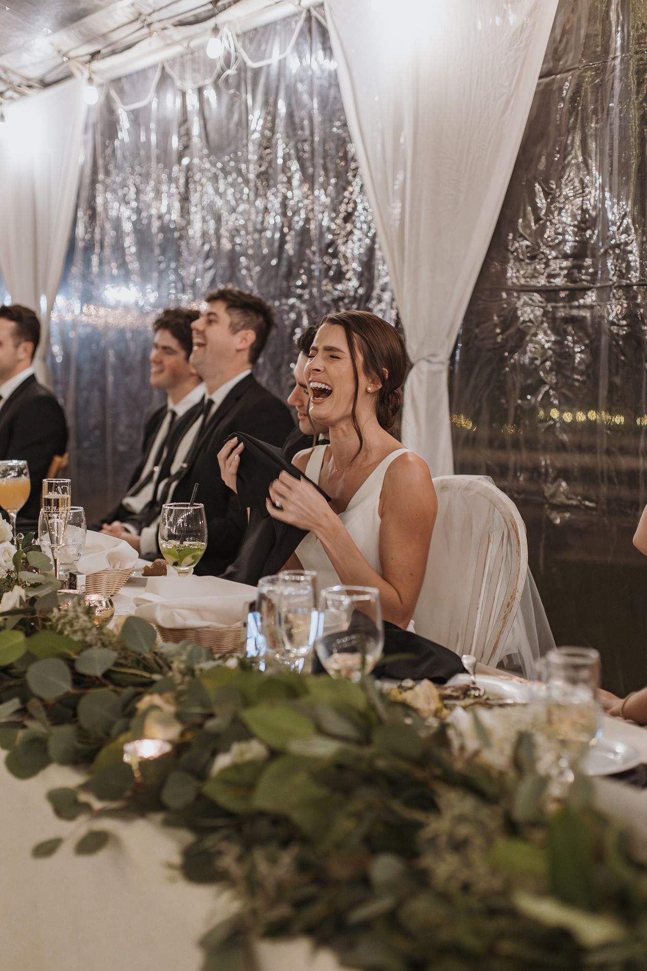 A bride laughs and enjoys themselves at an elegant wedding reception under a white tent with twinkling lights and greenery decor.