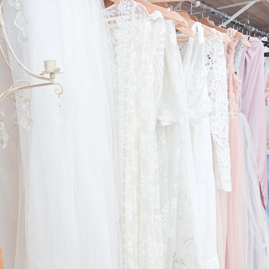 Wedding dresses and formal gowns in white lace and pastel colors hanging on a clothing rack in a bridal boutique.