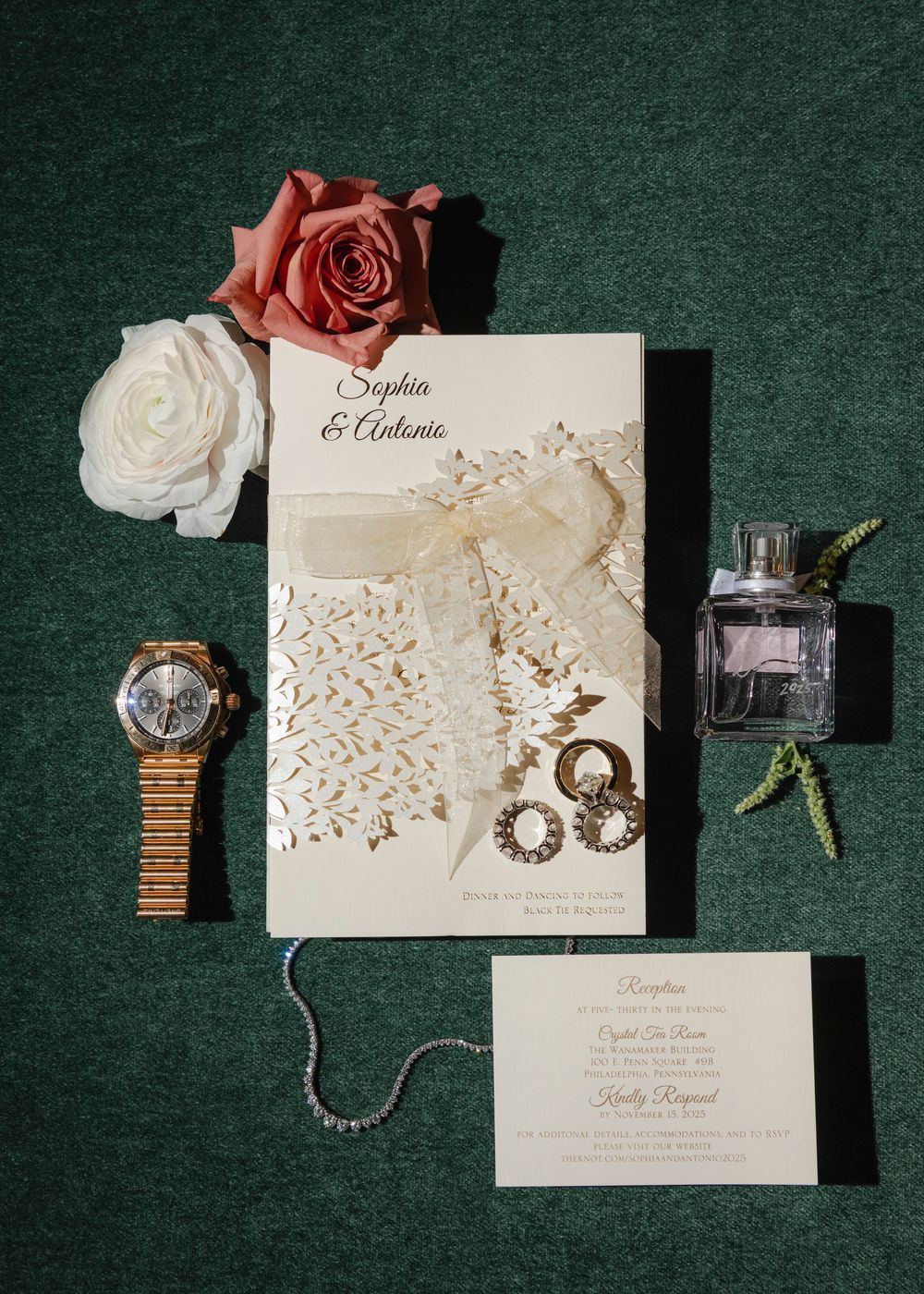 Wedding invitation suite with roses, jewelry, and perfume arranged on dark green velvet background.