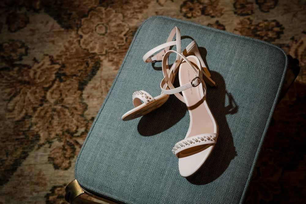 Elegant white bridal sandals with crystal embellishments resting on blue-gray fabric stool in moody lighting.