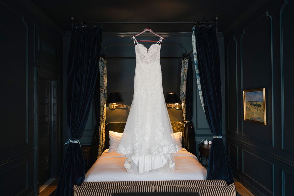 Elegant white wedding dress hanging in a dark fabric and wooden bed frame against moody lighting.