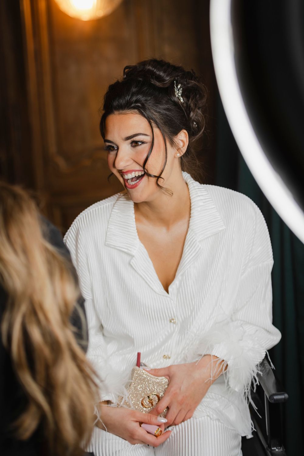 A candid moment of the bride wearing a white feathered pajama set laughing while waiting for the makeup artist