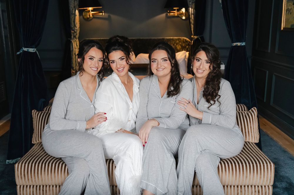 A bride and her 3 bridesmaids wearing light colored clothing laugh and smile together on a striped couch.