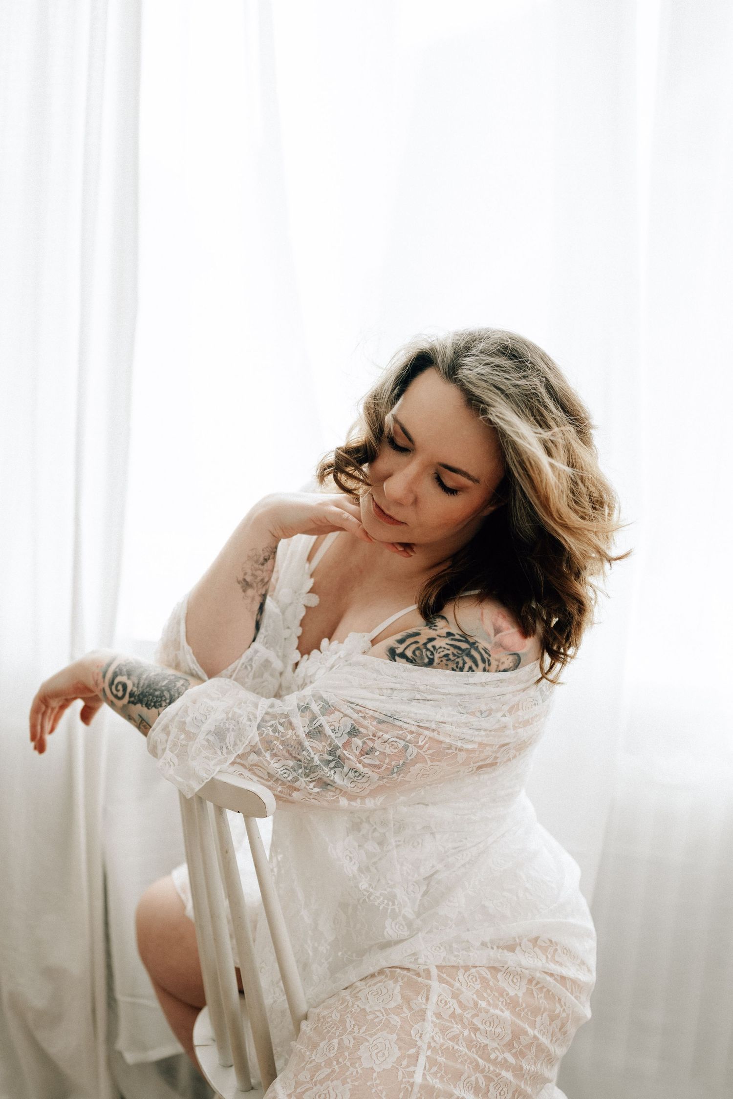 Boudoir Portrait einer Frau in einem Spitzencardigan sitzend auf einem weißen Stuhl am Fenster mit sanftem Licht.