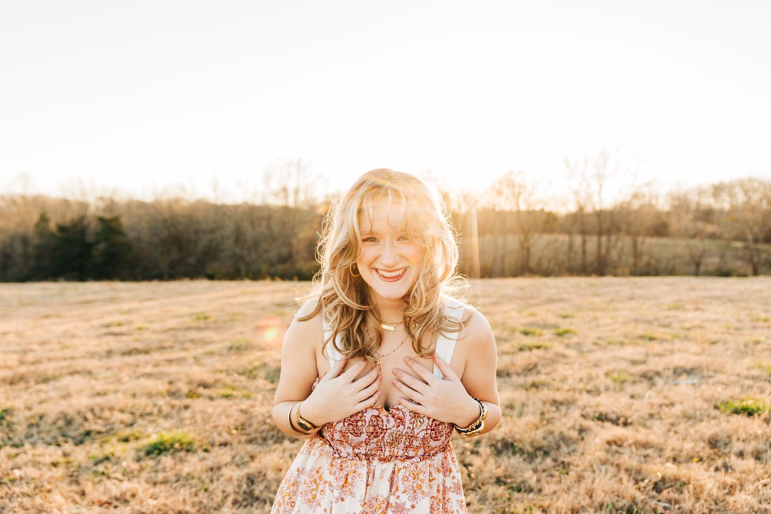 Close-up portrait of someone with wavy blonde hair wearing a floral top and layered gold necklaces, smiling outdoors.