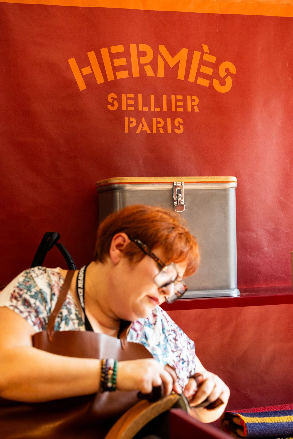 A woman works on leather goods beneath a red Hermes Sellier Paris sign.