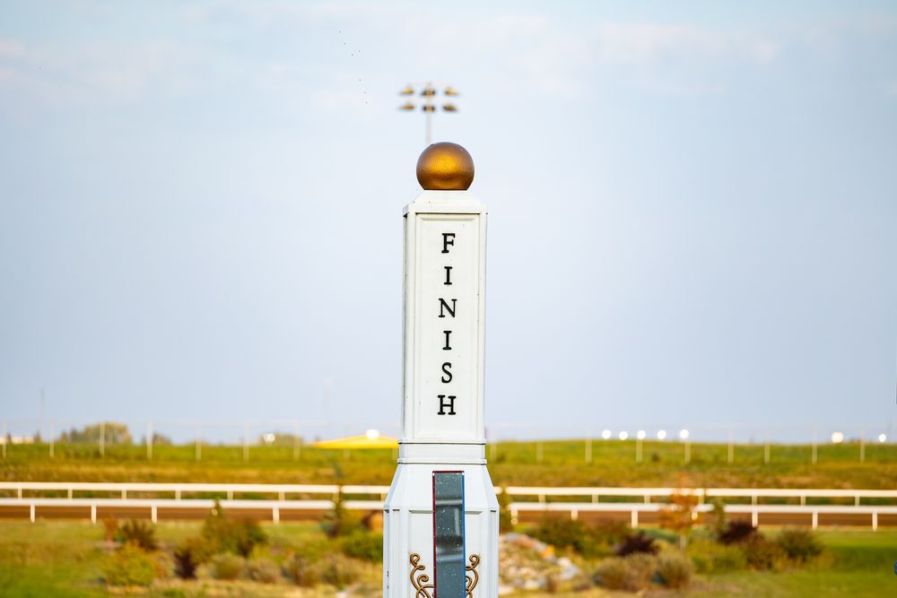 A white finish line post stands on a horse racing track under a cloudy sky.