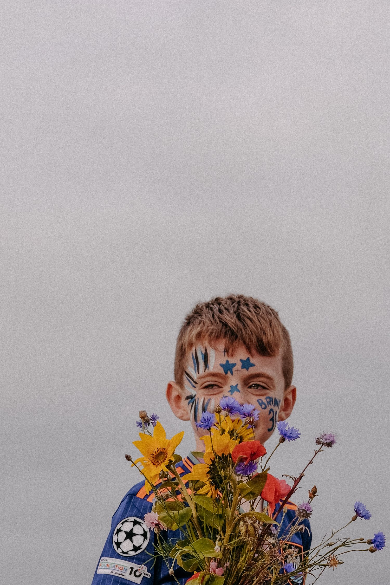 A young child playfully hiding their face behind a vibrant bouquet of wildflowers, captured in a bright and joyful lifestyle portrait.