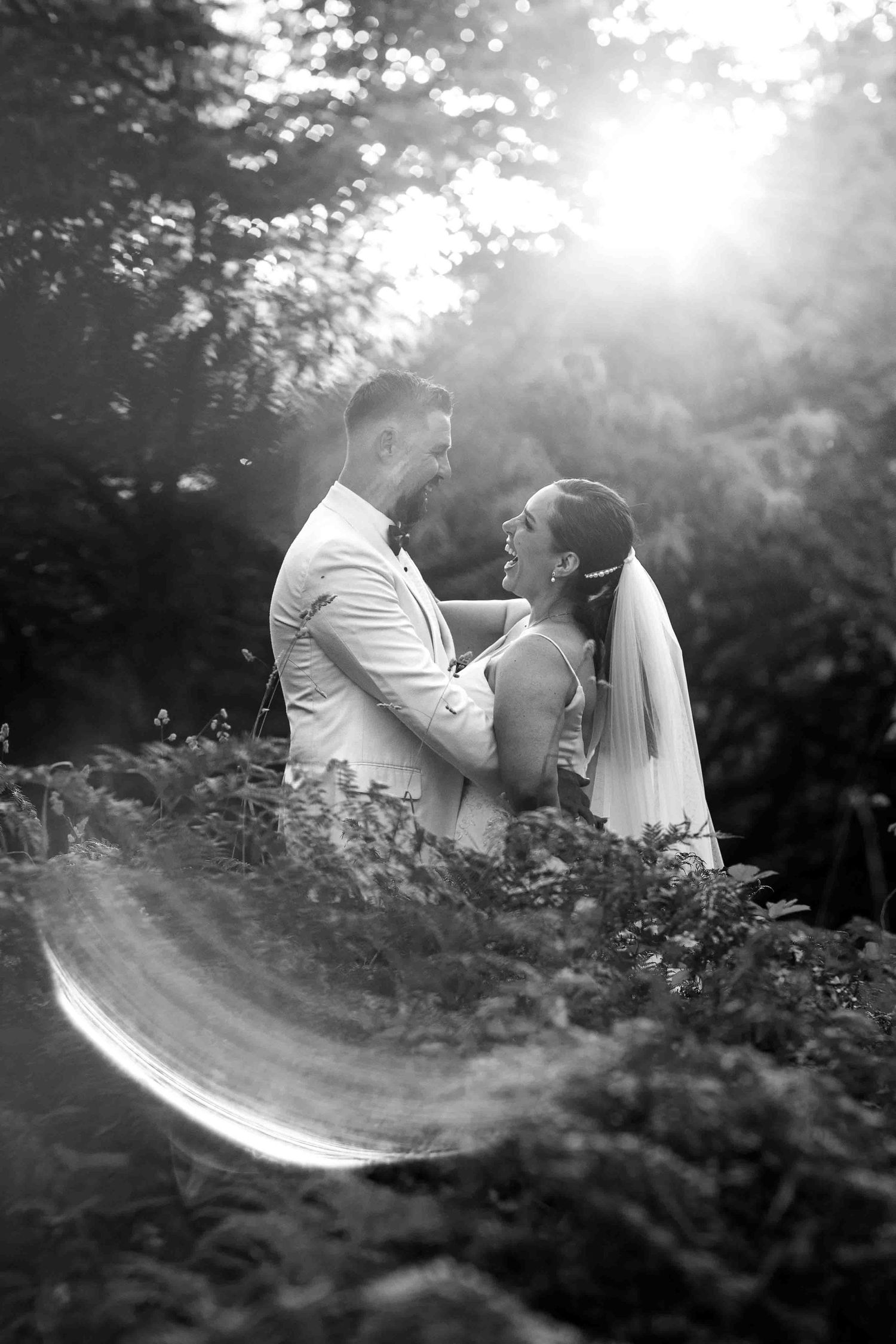 A bride and groom share an intimate moment in a sunlit garden setting captured in black and white photography.