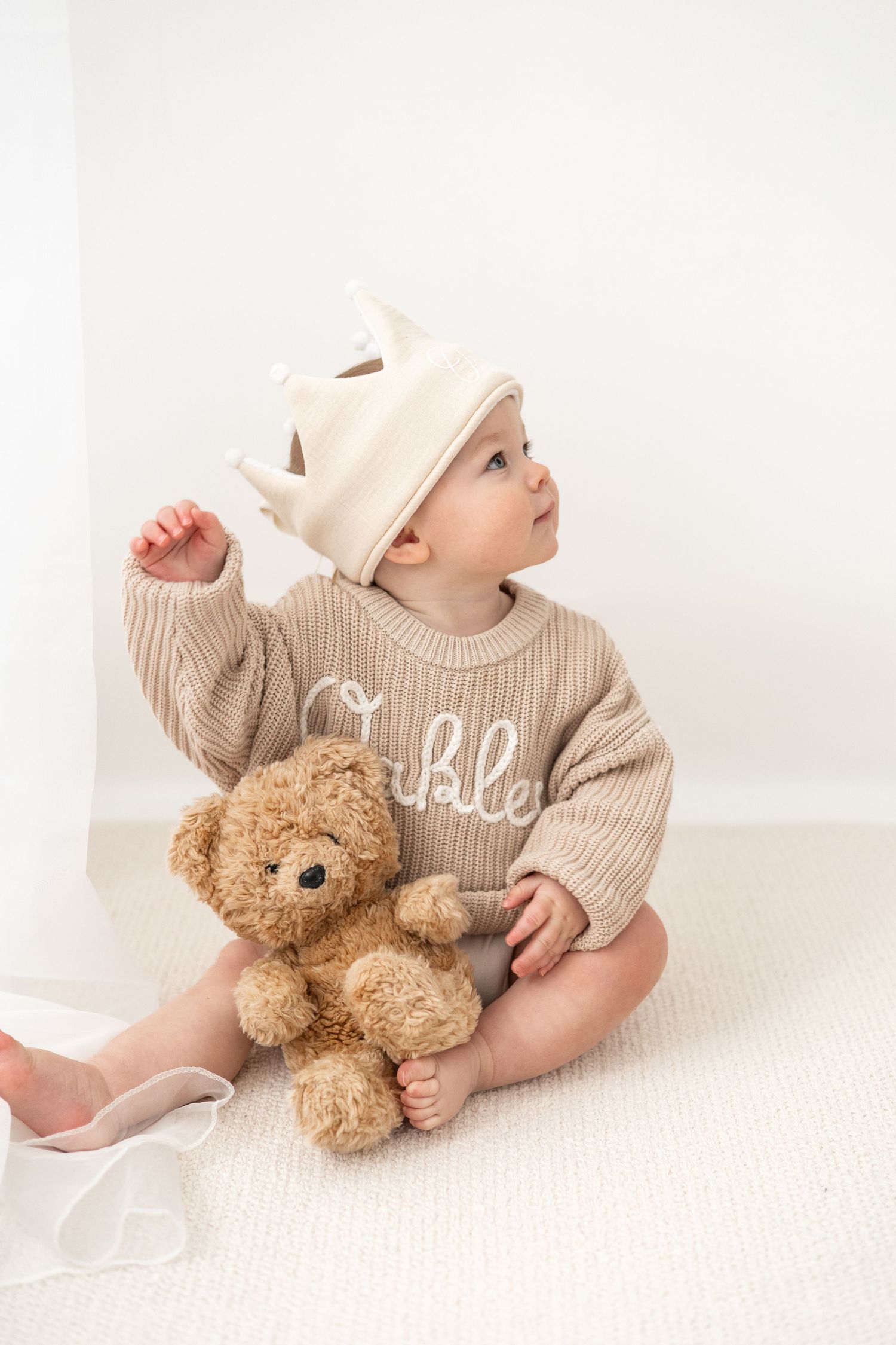 Baby wearing beige sweater and one crown sits with teddy bear on white background during Birthday shoot based in Nottingham