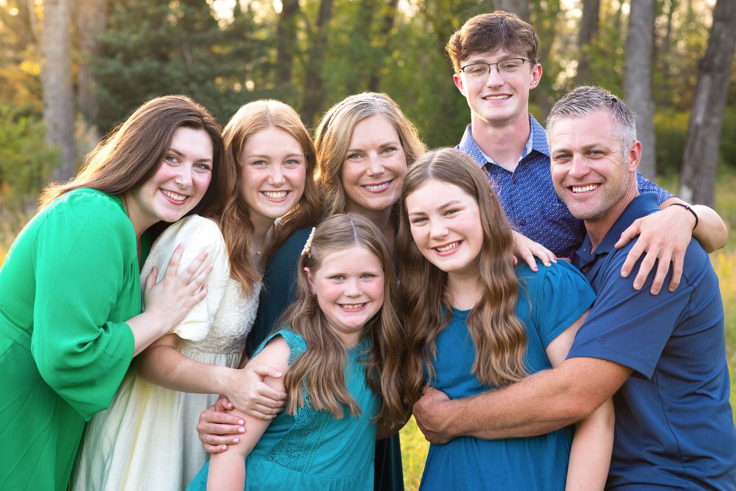 A family wearing blue and green shirts poses together outdoors during golden hour lighting.
