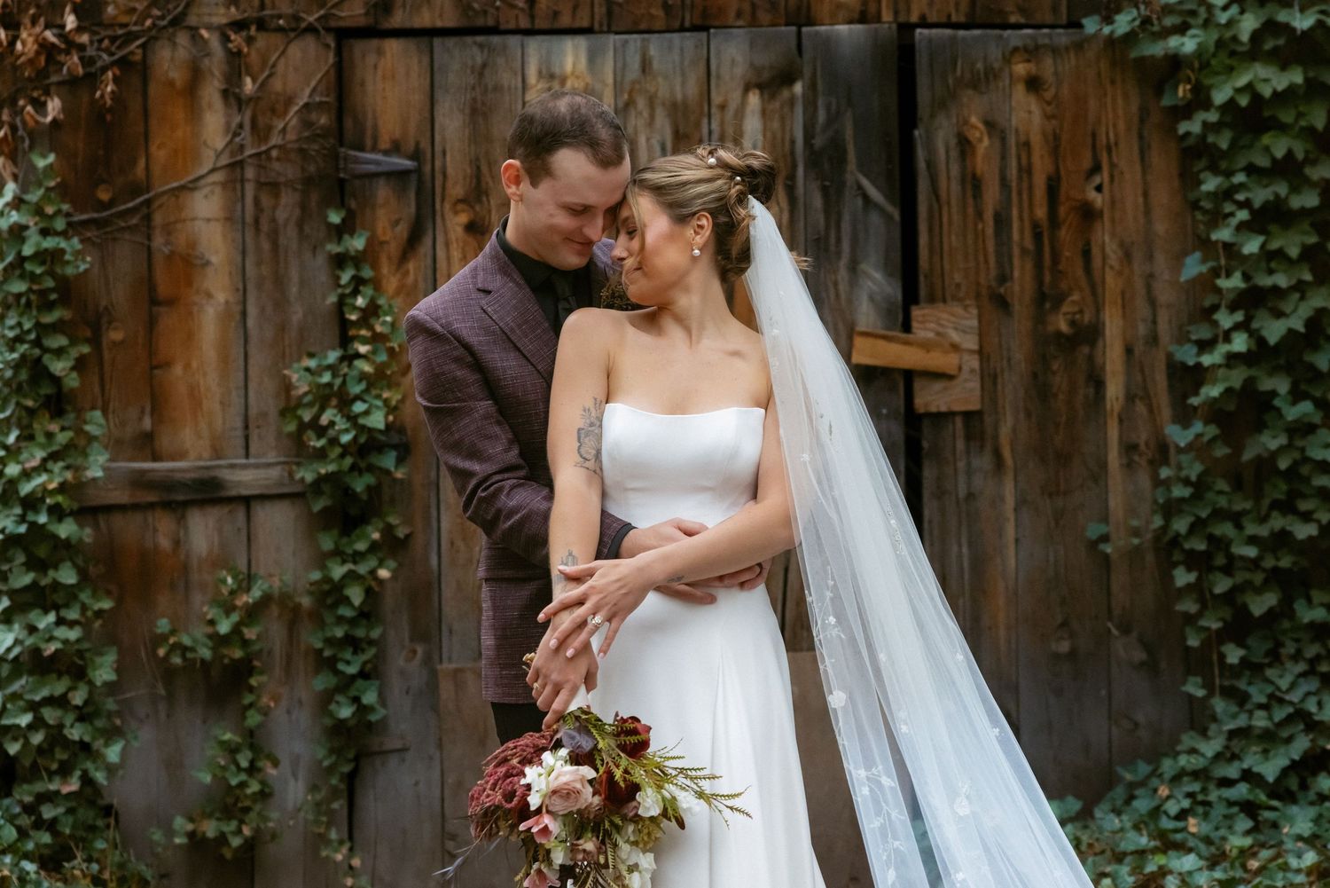 A couple shares an intimate moment in front of a rustic barn wall with climbing ivy, the bride holding an autumn bouquet.