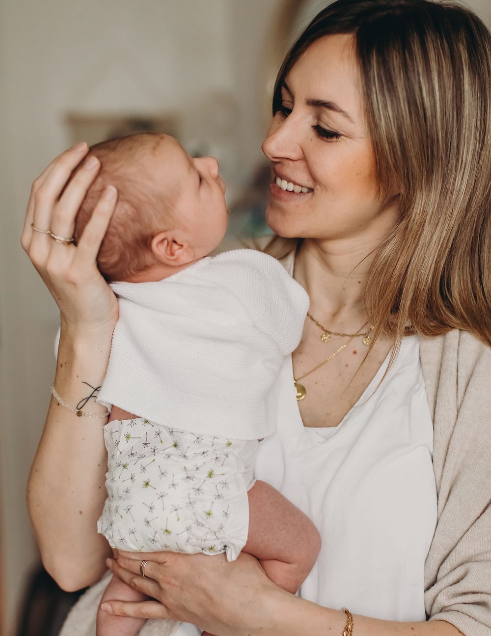 A mother in white and beige clothing smiles lovingly while holding and gazing at her newborn baby in a floral onesie.