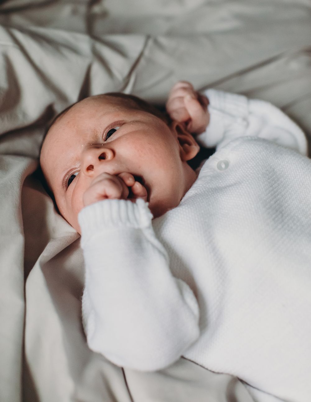 A newborn baby in a white sweater lies on white bedding in a series of candid close-up shots.