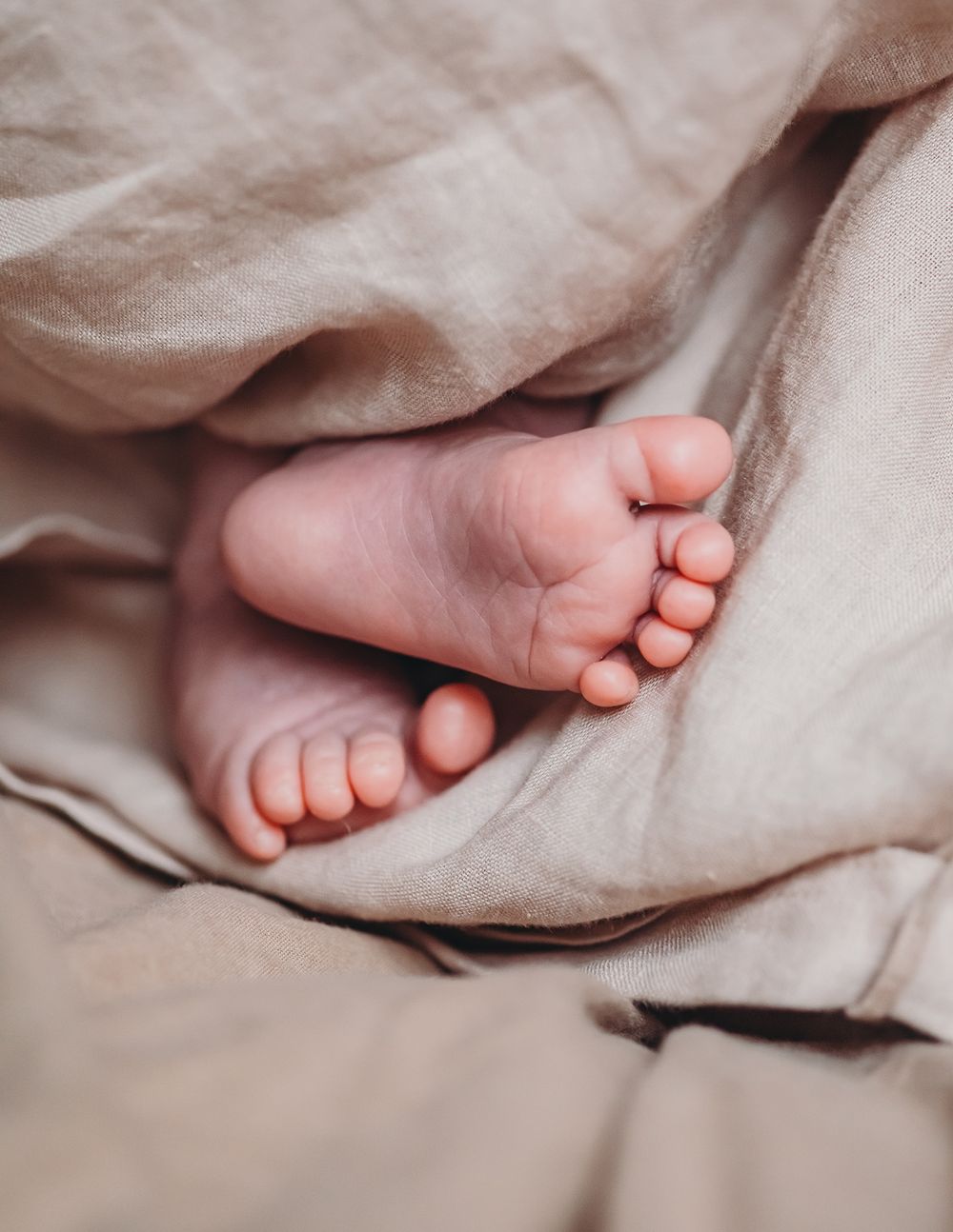 Tiny baby feet peek out from soft beige blankets in a gentle, intimate close-up photograph.