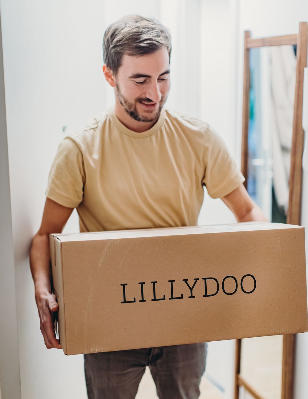 Person in beige shirt carrying large brown cardboard box with Lillydoo text through doorway while moving.