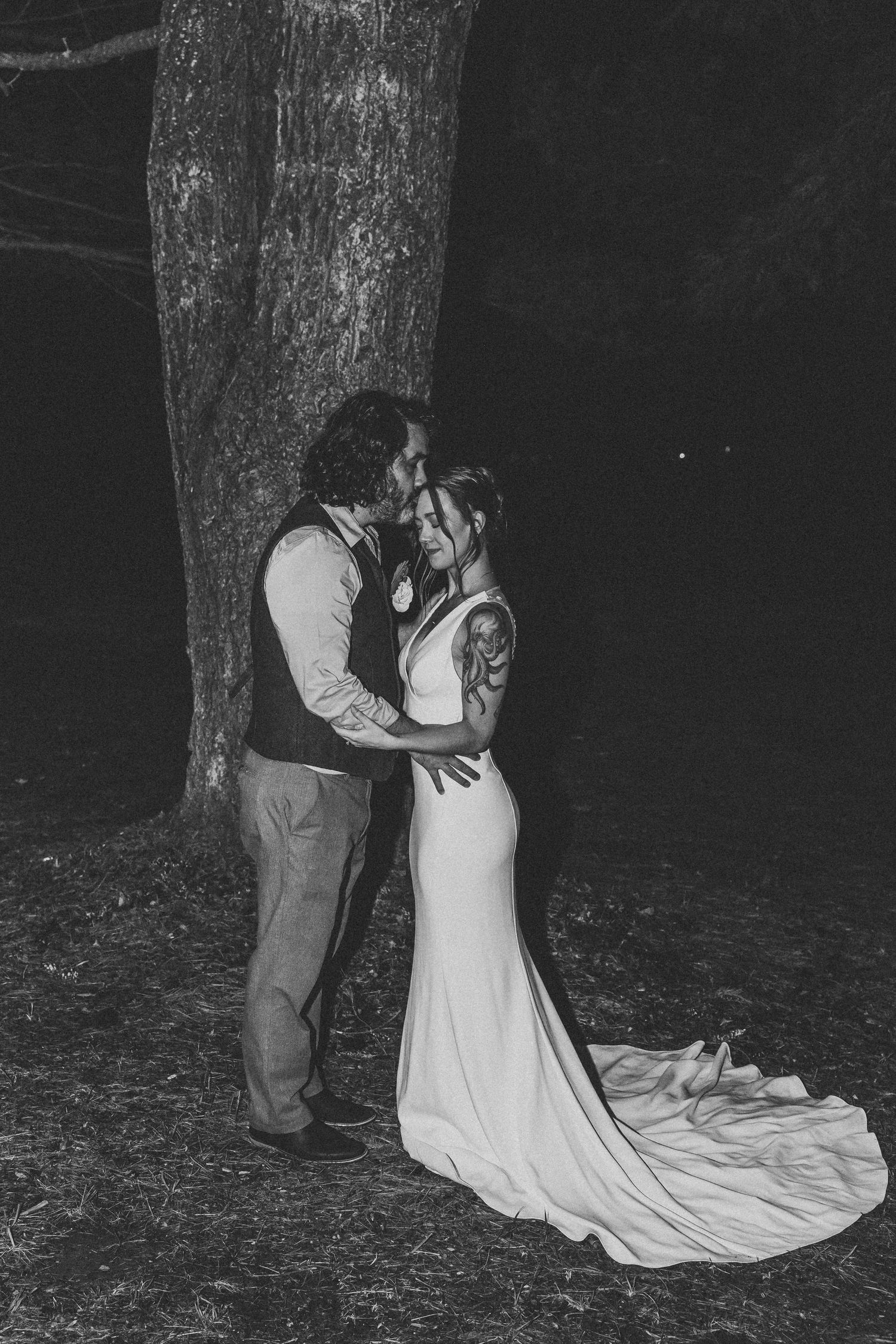 Black and white photo of bride and groom sharing romantic moment under tree at night.