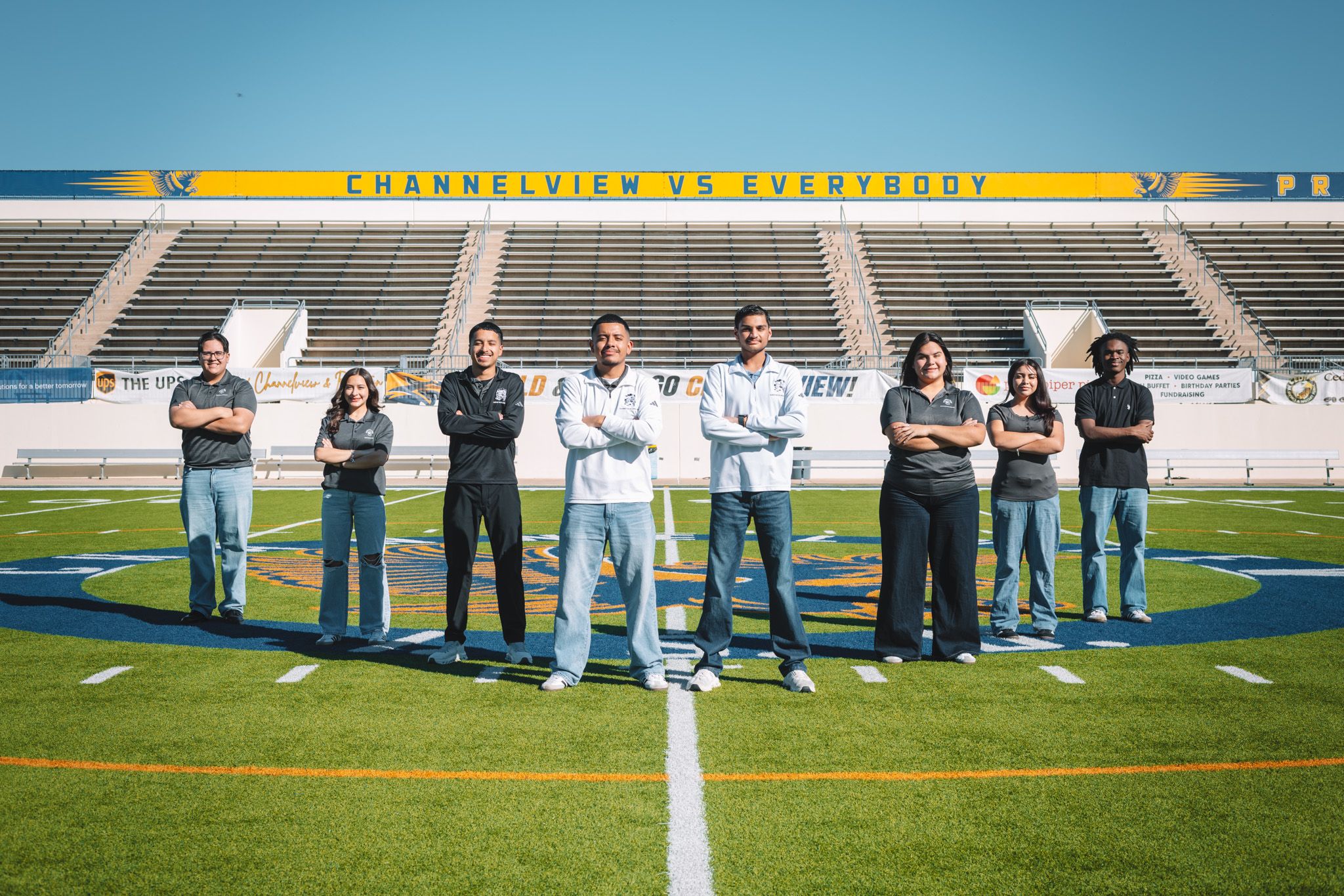 A group of people wearing matching light blue pants stand in formation on a football field with stadium seating behind them.