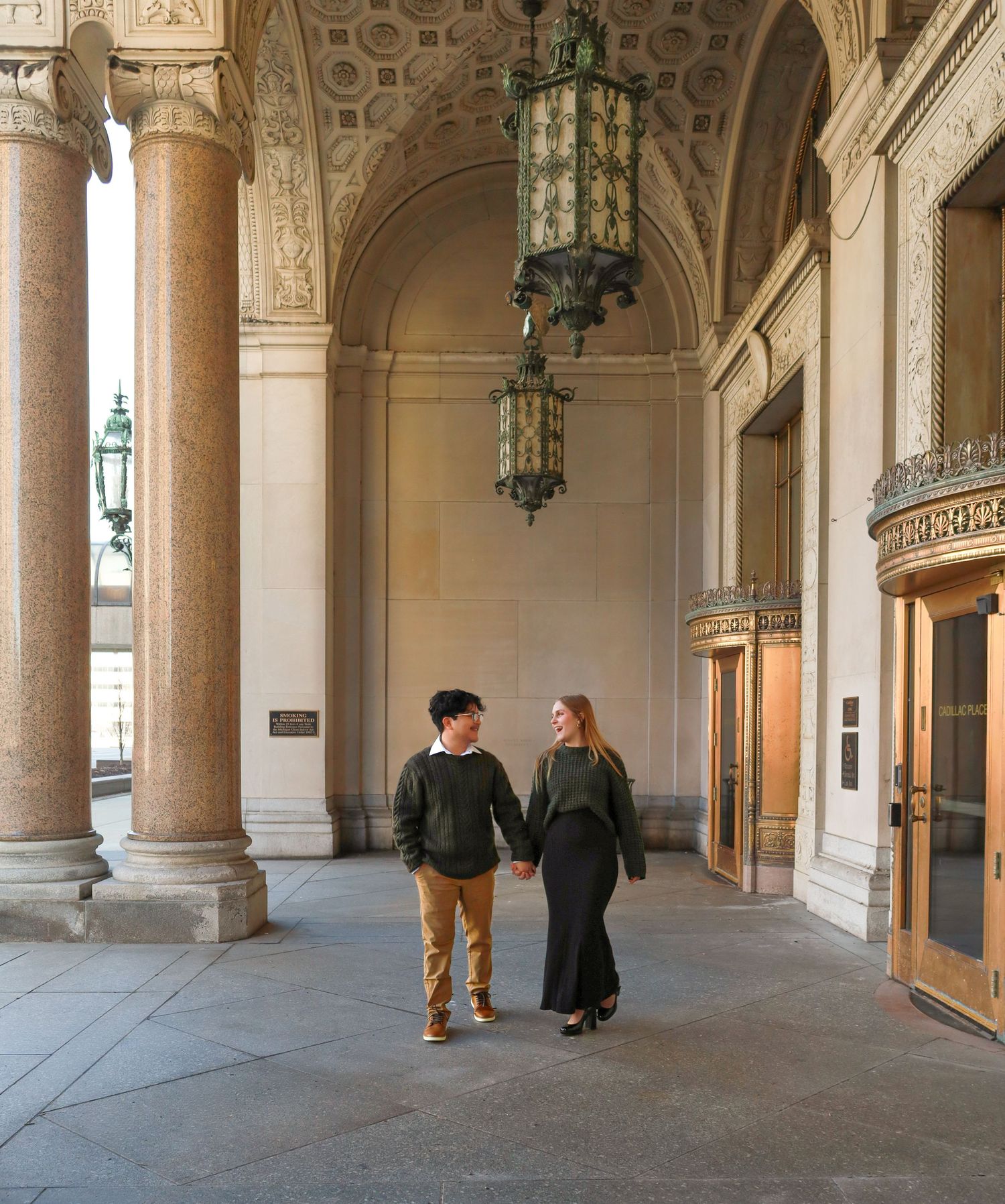 A couple walks together through an elegant colonnade with ornate architecture and hanging lanterns.