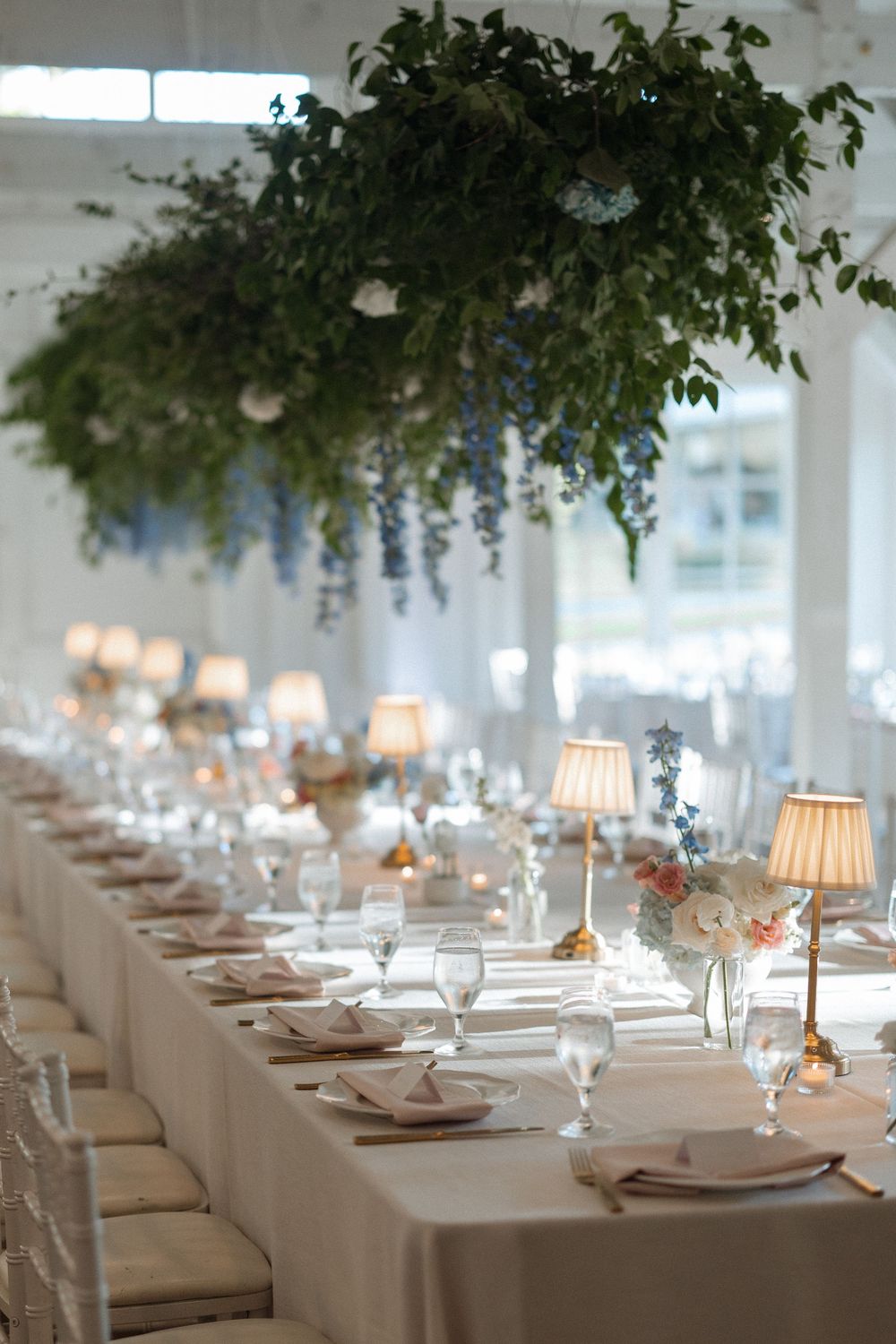 Elegant wedding reception tables with white linens, hanging greenery and soft lamp lighting create an intimate atmosphere.