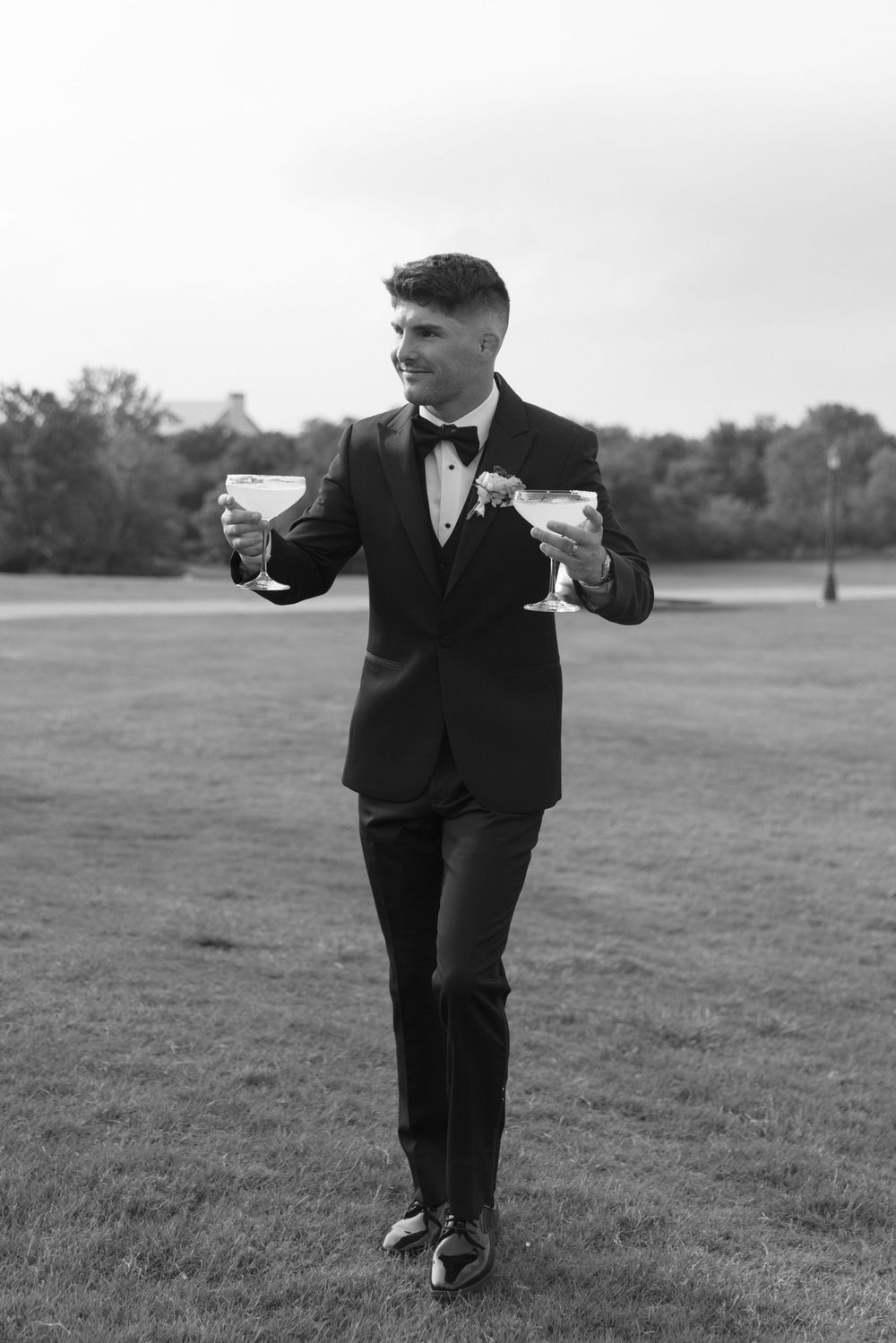 Groom in black tuxedo holding two champagne glasses outdoors on green lawn in black and white photo.