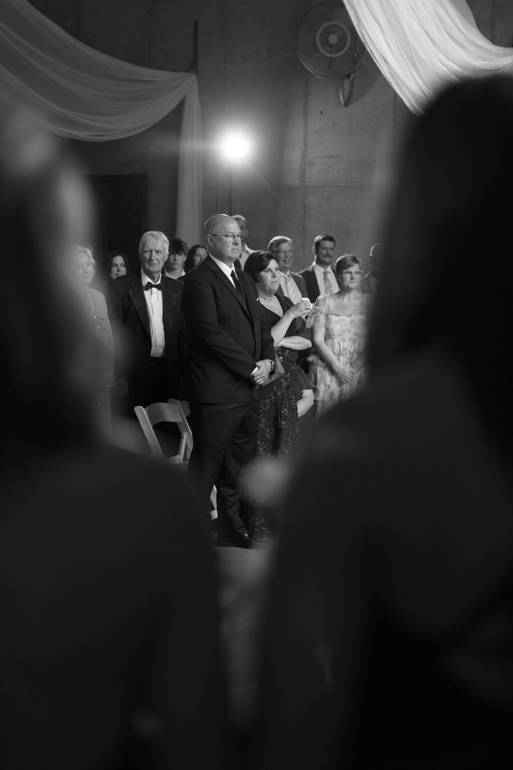 Black and white photo of elegantly dressed wedding guests standing and watching a ceremony in a grand venue.
