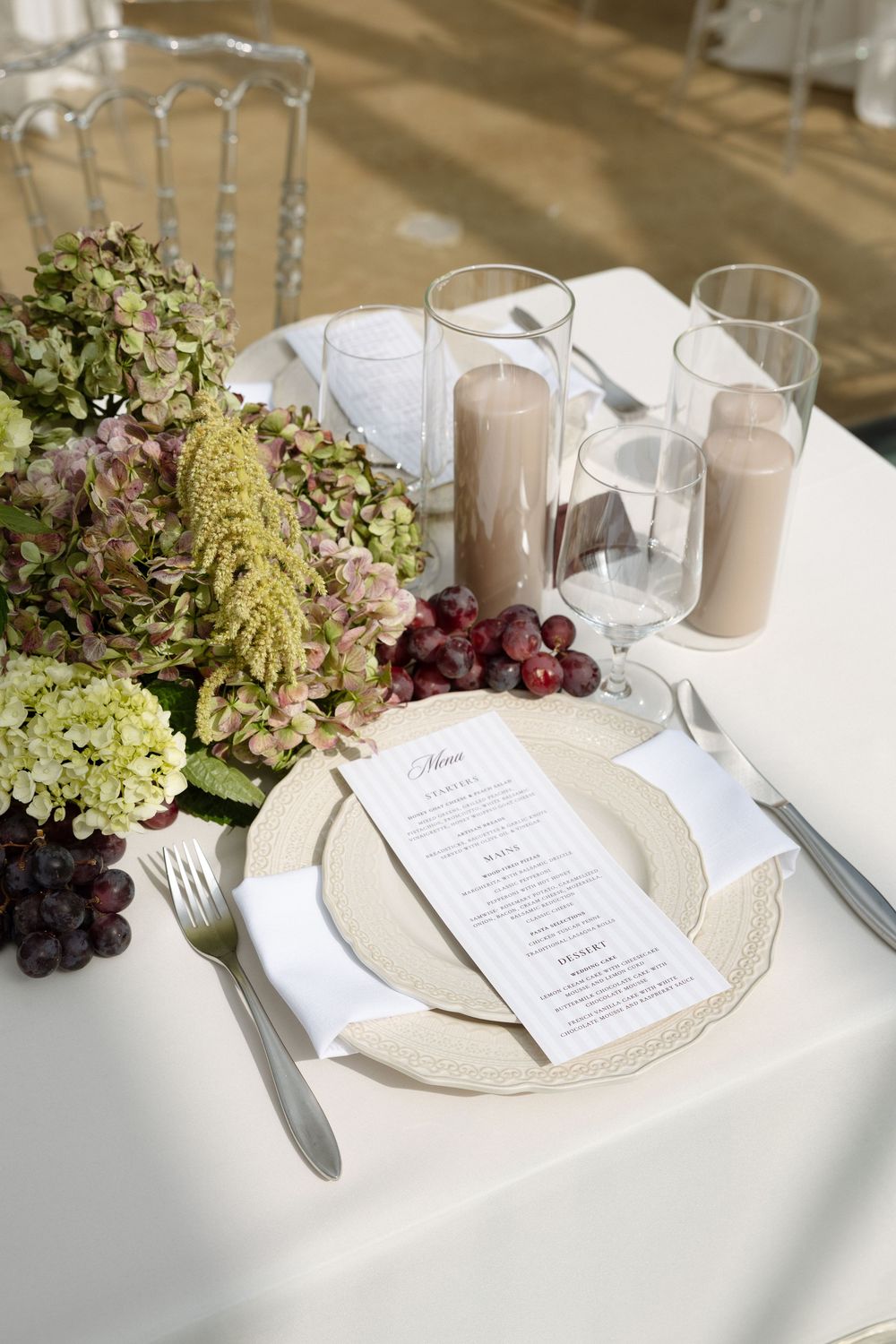 Elegant wedding table setting with hydrangeas, grapes, candles, and a menu card on a white tablecloth.