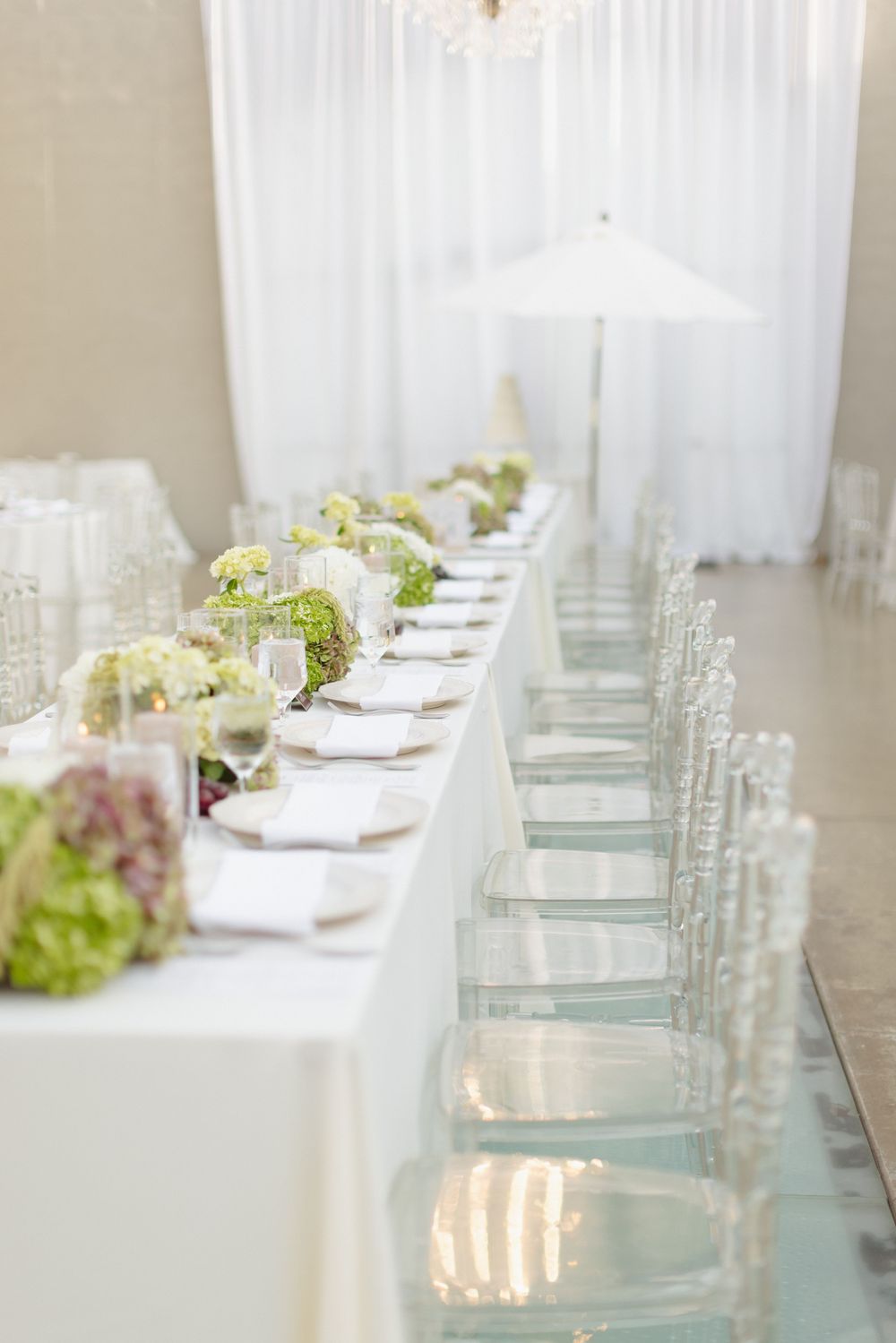 Elegant white wedding reception table with ghost chairs, green floral centerpieces, crystal glassware, and sheer draping backdrop.