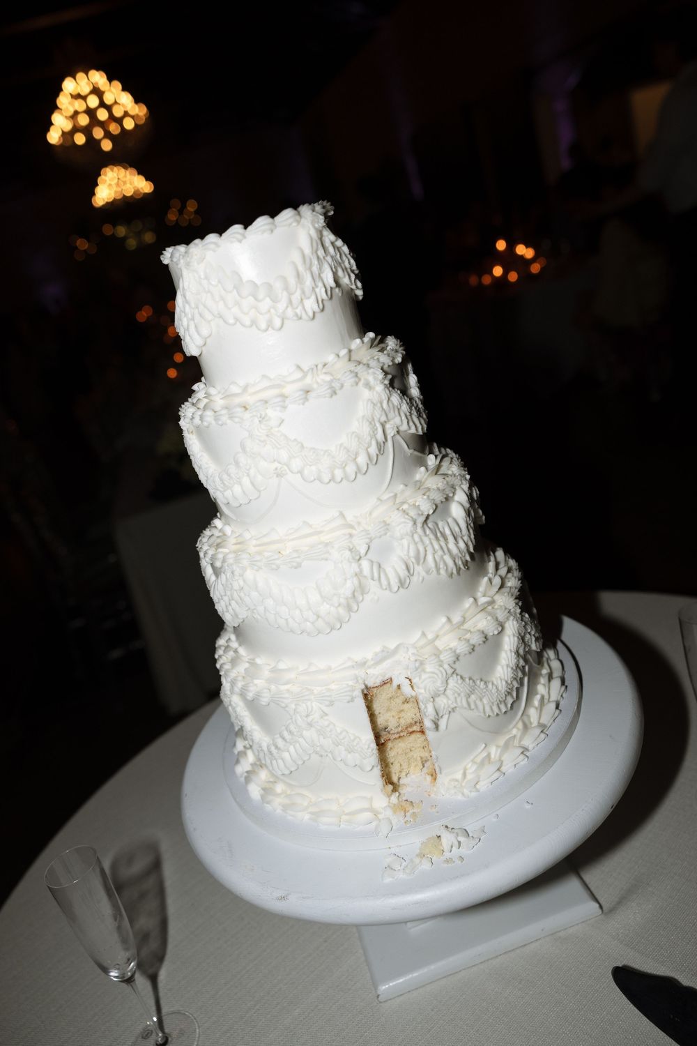 Elegant four-tier white wedding cake with rope frosting details on a white stand, partially cut, with bokeh lights in background.
