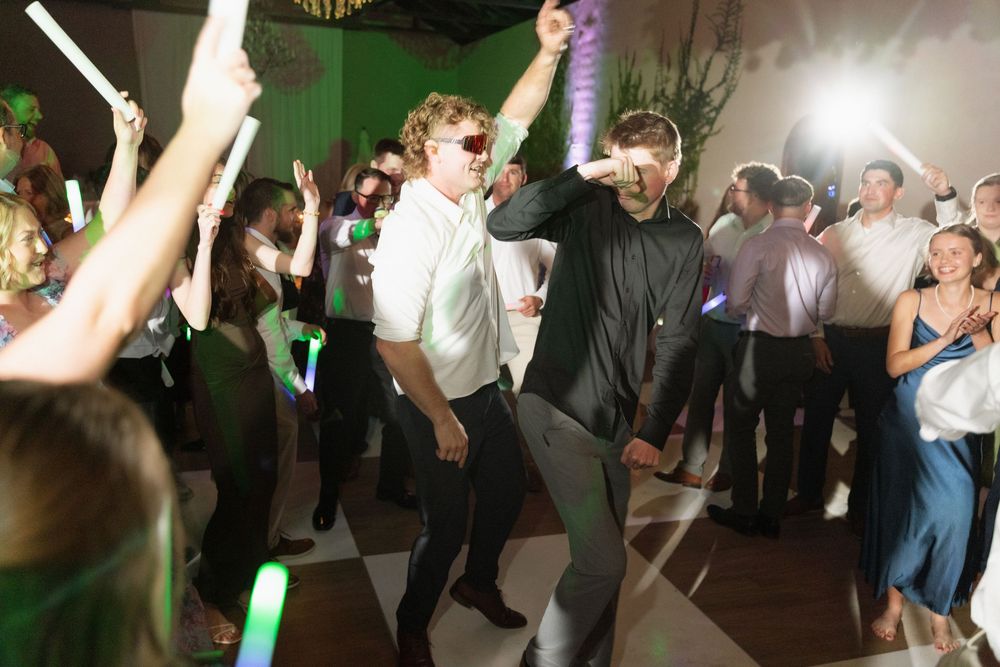 Guests dancing energetically at a wedding reception, holding glow sticks under bright lights on a crowded dance floor.