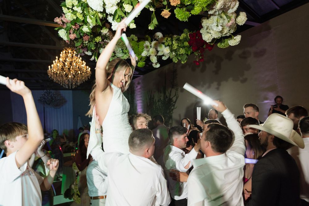 Bride lifted by guests on crowded wedding dance floor with glowsticks, floral chandelier, and chandelier lights above.