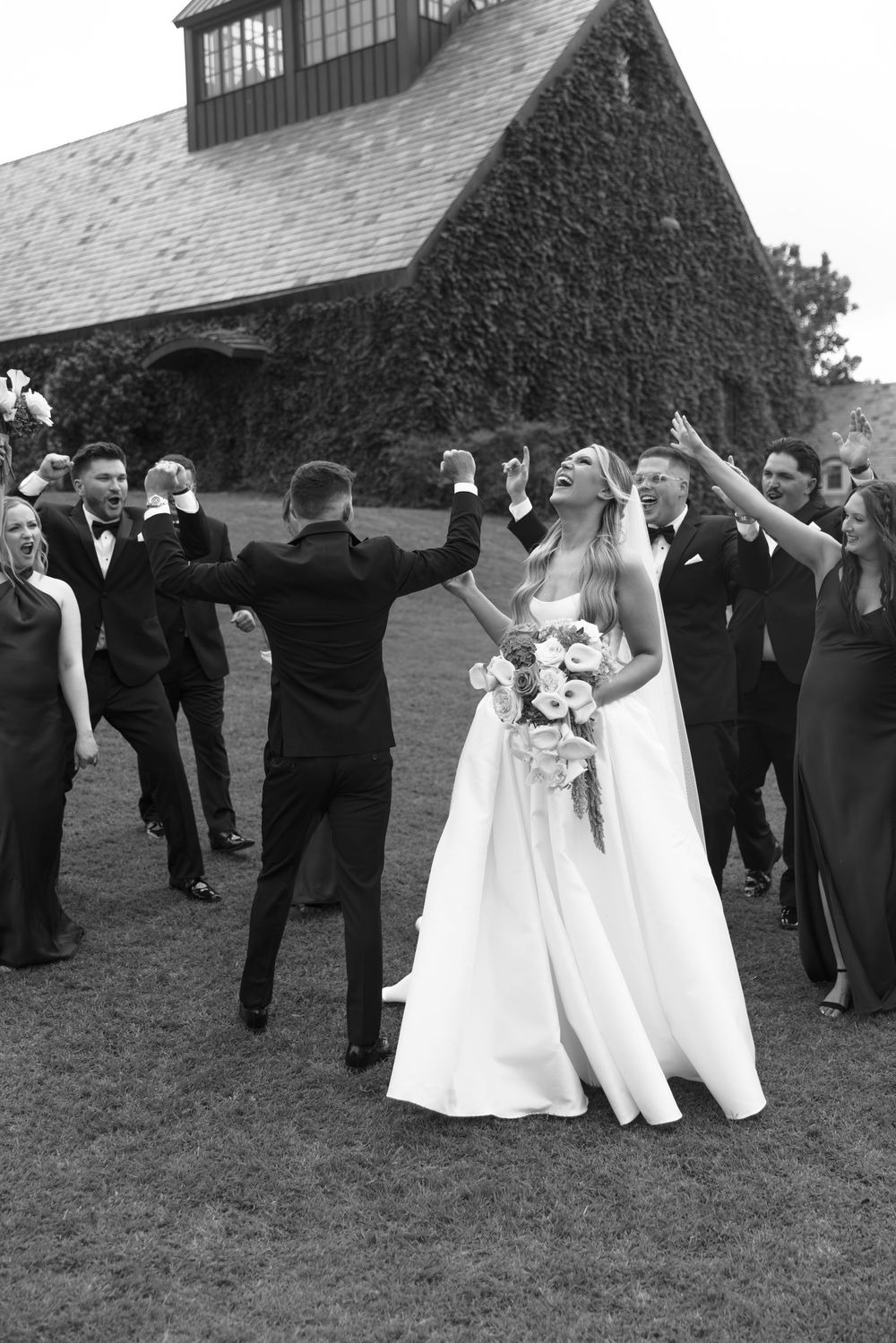 Black and white photo of a joyful bride in a white gown celebrating with wedding party outside a stone building.