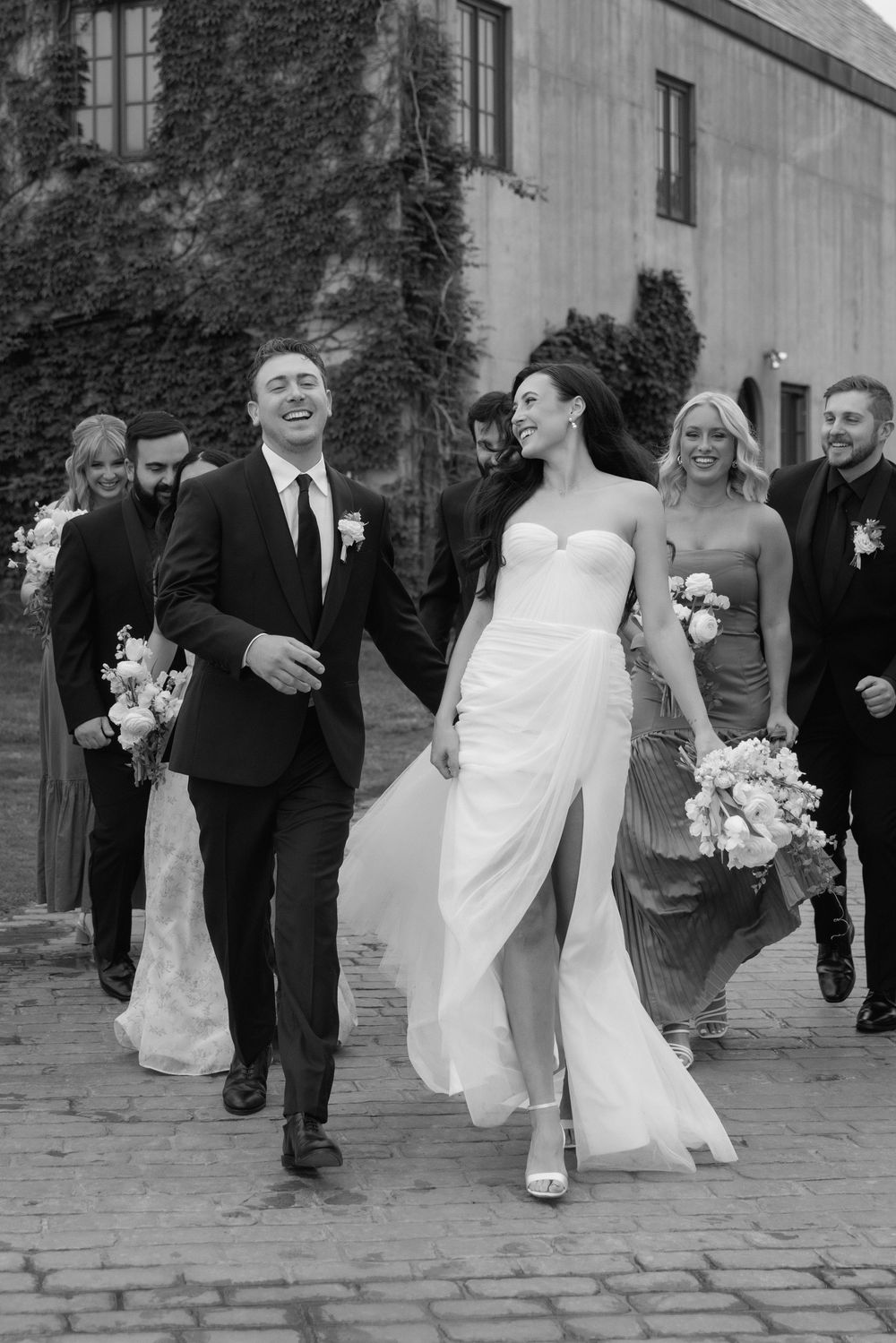 A wedding party walks along a cobblestone path in black and white, with the bride's white dress flowing in motion.