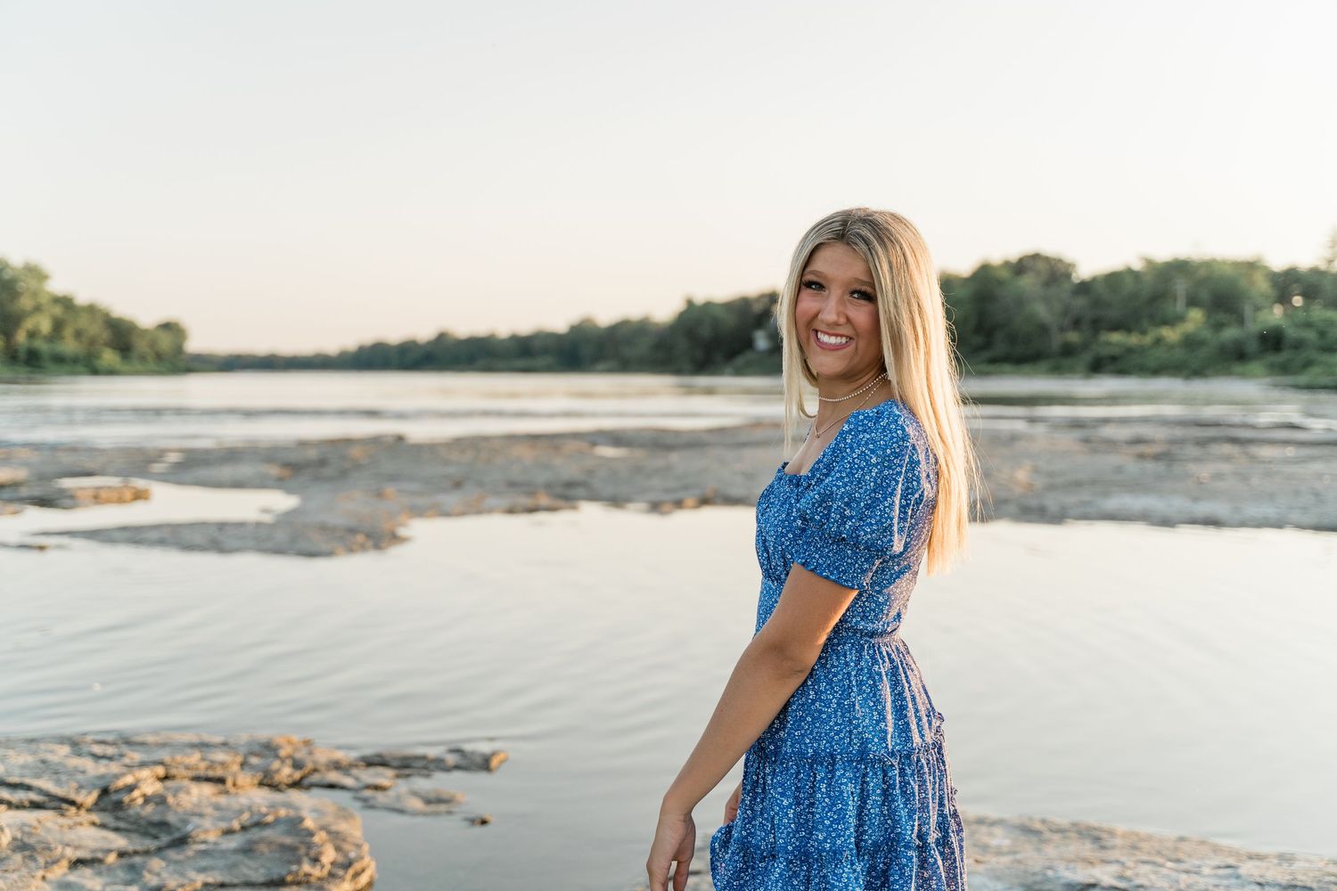 Perrysburg senior in blue floral dress poses by a scenic riverside during golden hour sunset at Side Cut Metropark